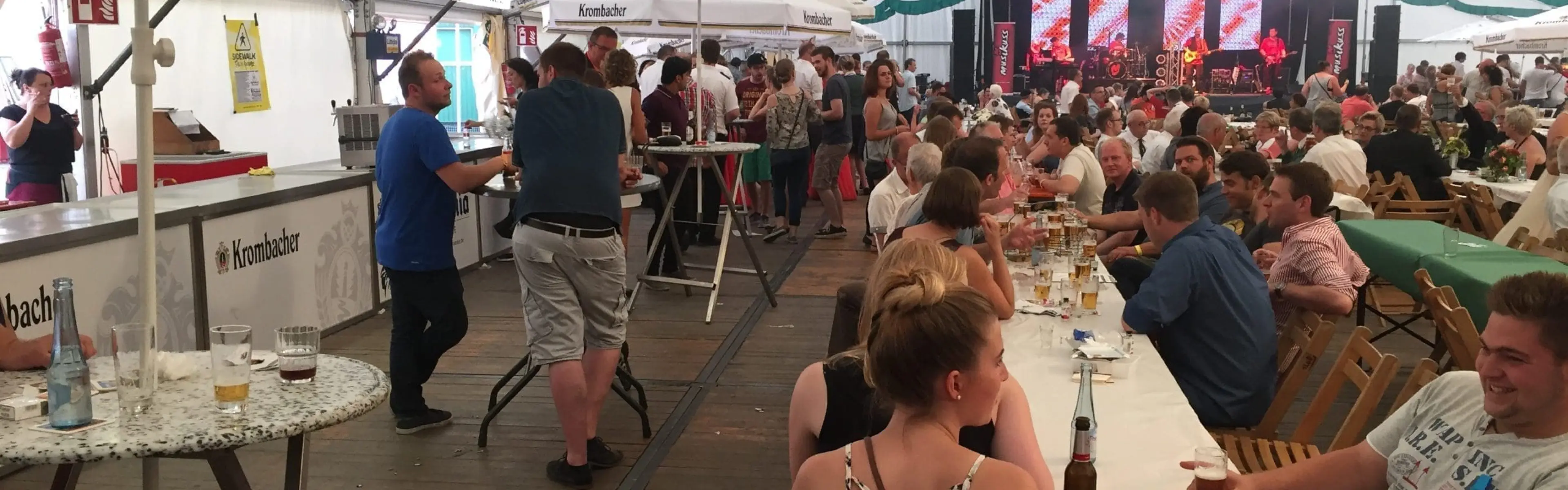 2023_Schuetzenfest_Zeltbestuhlung MontagFrühschoppenFoto1 Die Menschen sitzen an langen Tischen in einem großen Zelt, trinken Bier und unterhalten sich. Einige stehen in der Nähe des Barbereichs. Das Zelt ist mit grünen Bannern geschmückt, ein Regenschirm wirbt für Krombacher, und im Hintergrund ist eine Bühne mit Lichtern zu sehen.