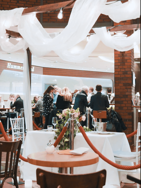 Hanseviertel_ValentinsDay Menschen versammeln sich und unterhalten sich um einen Esstisch in einem stilvollen, überdachten Restaurant mit drapiertem weißen Stoff über dem Kopf. Leere, gedeckte Tische mit Blumen und Weinkübeln stehen im Vordergrund, getrennt durch ein rotes Seil.