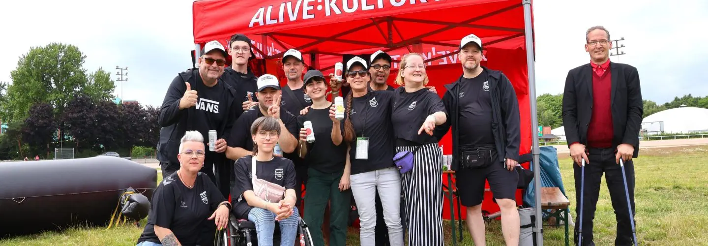 IMG_4571 Eine Gruppe von Menschen posiert für ein Foto vor einem roten Zelt mit der Aufschrift ALIVE KULTUR e.V. auf einer Wiese. Die meisten tragen schwarze Hemden und Mützen. Eine Person sitzt im Rollstuhl, eine andere steht mit einem Gehstock.