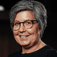 Nicole Eine Person mittleren Alters mit kurzen grauen Haaren, die eine Brille und ein dunkles Oberteil trägt, lächelt in die Kamera vor einem dunklen Hintergrund.