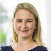 Selina Schumacher Eine Frau mit glattem blondem Haar, die ein marineblaues Oberteil und Perlenohrringe trägt, lächelt in einer hell erleuchteten Innenumgebung in die Kamera.