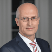 tschentscher Ein Mann mittleren Alters mit Brille, kurzen grauen Haaren und glatt rasiertem Gesicht trägt einen dunklen Anzug, ein weißes Hemd und eine gestreifte Krawatte und posiert vor einem unscharfen Innenhintergrund.