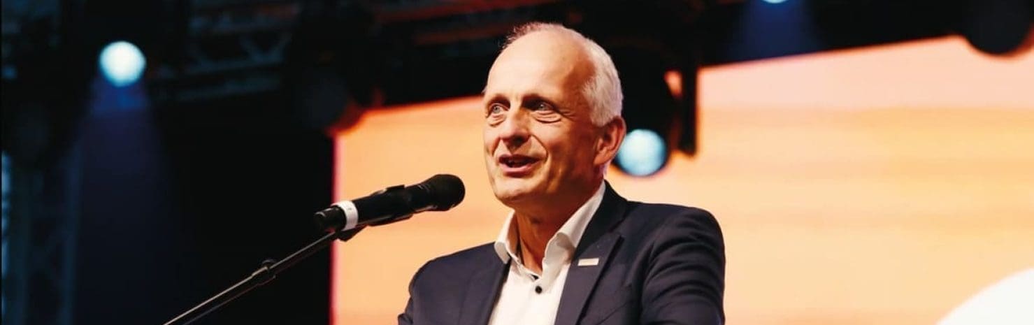 Ein Mann im Anzug spricht auf der Bühne in ein Mikrofon, während hinter ihm Bühnenlicht und ein orangefarbener Hintergrund zu sehen sind.