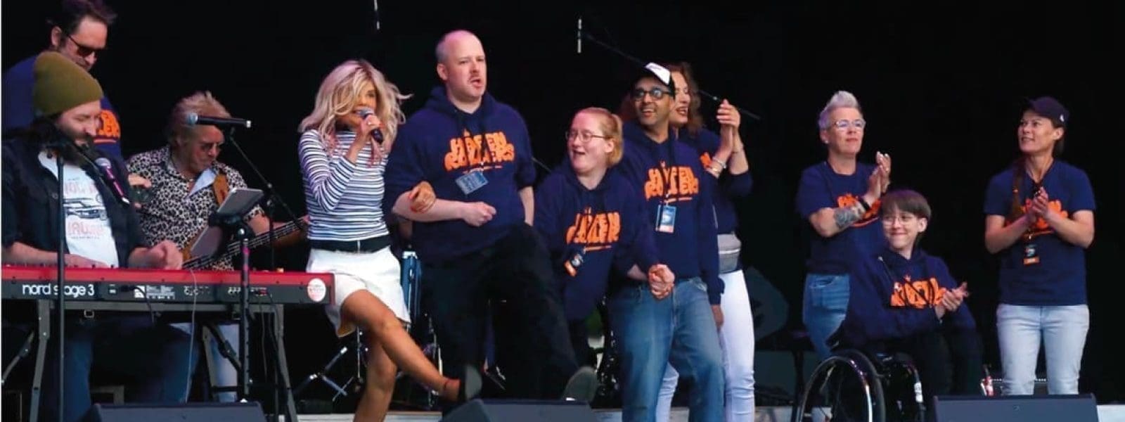 Eine Gruppe von Menschen, einige in passenden marineblauen G.I.V.E.R.-Hemden, steht und sitzt auf der Bühne. Eine Frau in der Mitte singt in ein Mikrofon, während andere lächeln, klatschen oder Rollstühle benutzen. Auf der linken Seite ist ein Keyboard zu sehen.