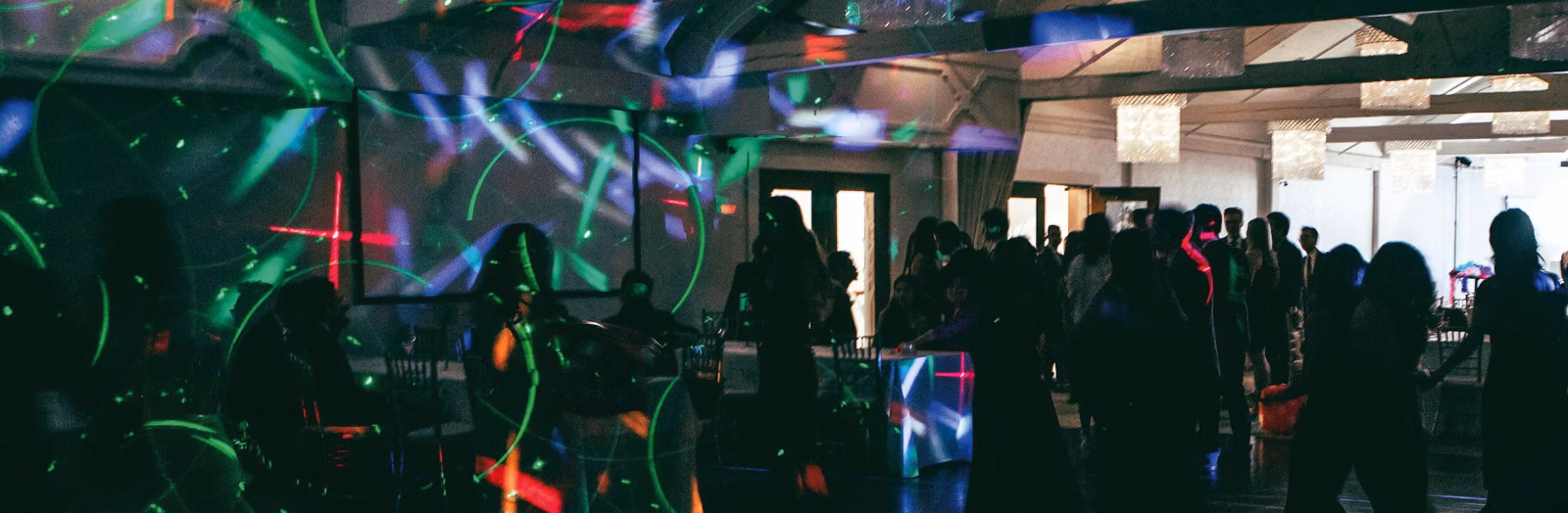 Die Menschen sind in einem schwach beleuchteten Raum versammelt, auf den grünes und rotes Laserlicht projiziert wird, wodurch eine Partyatmosphäre entsteht. Einige Gäste sitzen, andere stehen oder tanzen an beleuchteten Tischen.