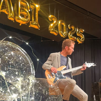 Zwei Musiker treten auf der Bühne auf, einer spielt Bassgitarre, der andere Akustikgitarre. Hinter ihnen befinden sich goldene Luftballons mit der Aufschrift ABI 2025, schwarze Vorhänge und große durchsichtige Ballons mit Lichterketten im Inneren.