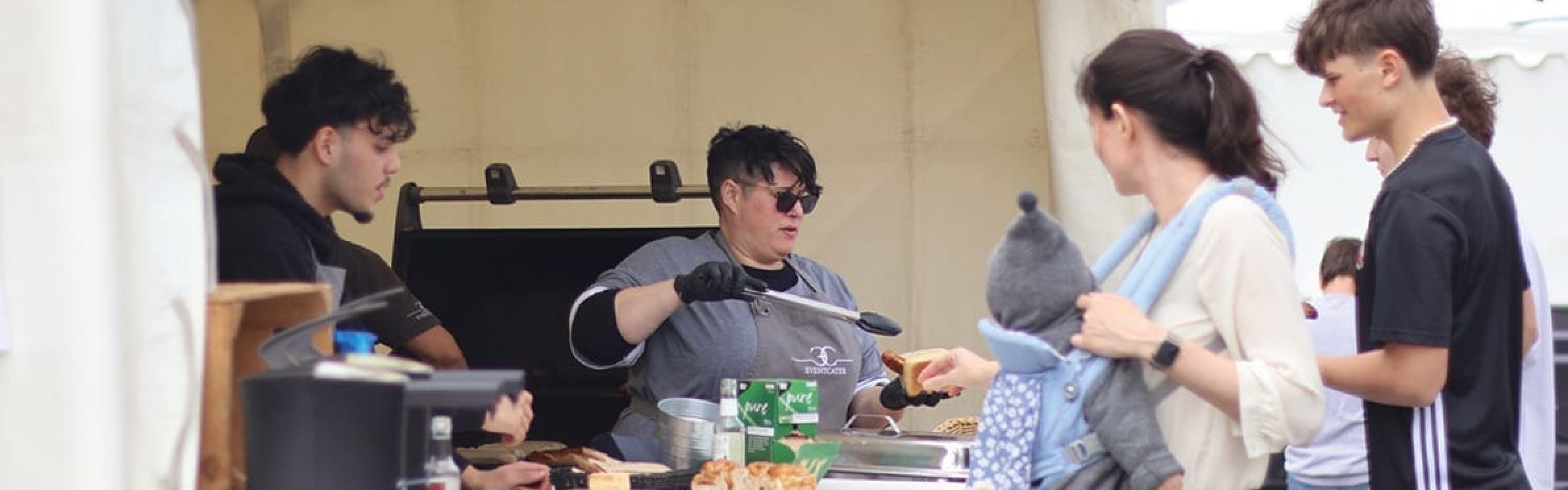 Eine Person mit schwarzen Handschuhen benutzt eine Zange, um das Essen an einem Stand im Freien zu servieren, während andere, darunter eine Frau mit einem Baby im Arm, in der Schlange warten. Ein weiterer Mitarbeiter und Lebensmittel sind auf dem Tresen zu sehen.