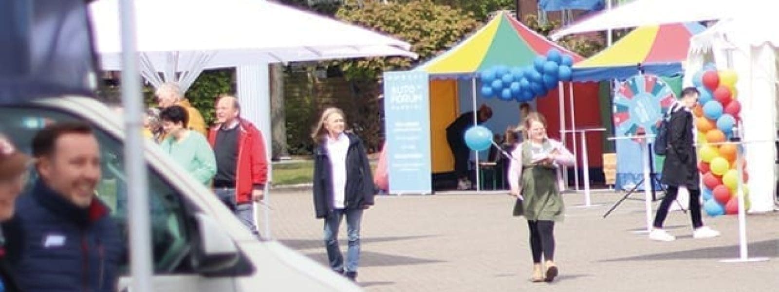 Menschen gehen und stehen bei einer Veranstaltung im Freien mit weißen Zelten, bunten Luftballons und Bannern im Hintergrund. Eine Frau hält Papiere in der Hand, während sie sich der Kamera nähert, und andere stehen in der Nähe eines Lieferwagens im Vordergrund.