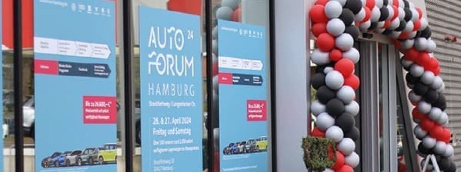 Ein gläserner Gebäudeeingang, geschmückt mit einem rot-weiß-schwarzen Luftballonbogen. Plakate an den Fenstern werben für das Auto Forum Hamburg mit Datum, Ort und Bildern von Autos.