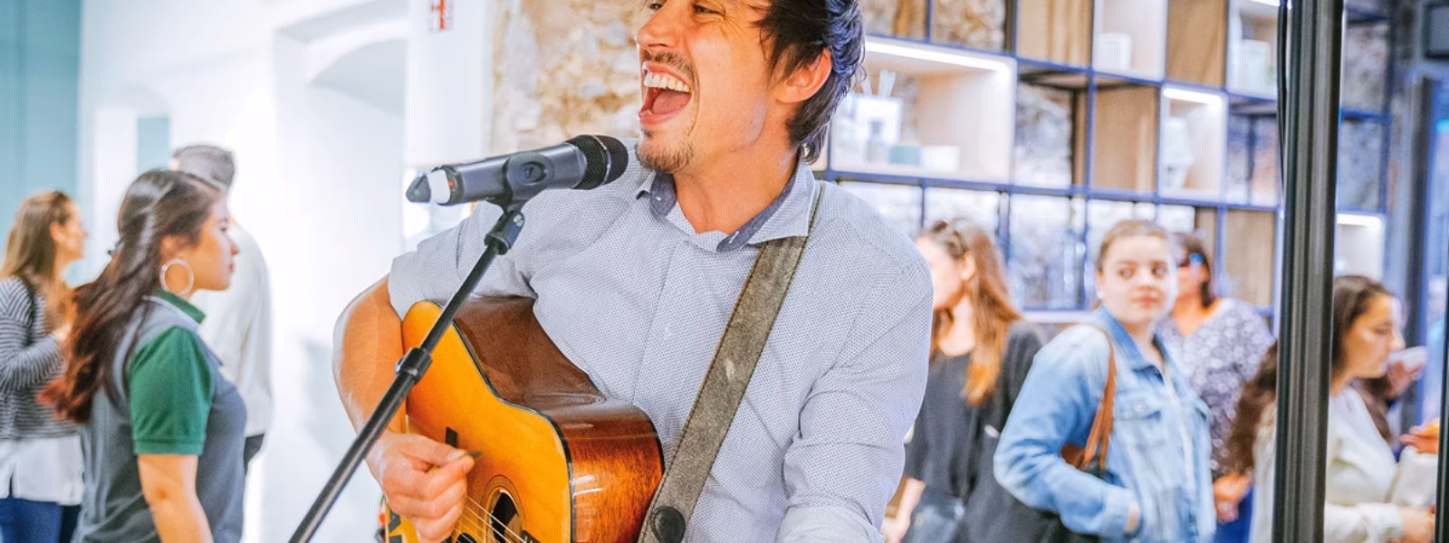 Ein Mann spielt eine Akustikgitarre und singt in ein Mikrofon bei einer Indoor-Veranstaltung. Im Hintergrund sind mehrere Personen in Freizeitkleidung zu sehen, von denen einige gehen und andere in der Nähe einer mit Regalen versehenen Wand stehen.