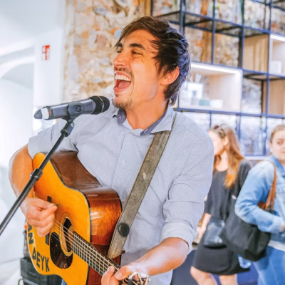 Ein Mann spielt eine Akustikgitarre und singt in ein Mikrofon bei einer Indoor-Veranstaltung. Im Hintergrund sind mehrere Personen in Freizeitkleidung zu sehen, von denen einige gehen und andere in der Nähe einer mit Regalen versehenen Wand stehen.
