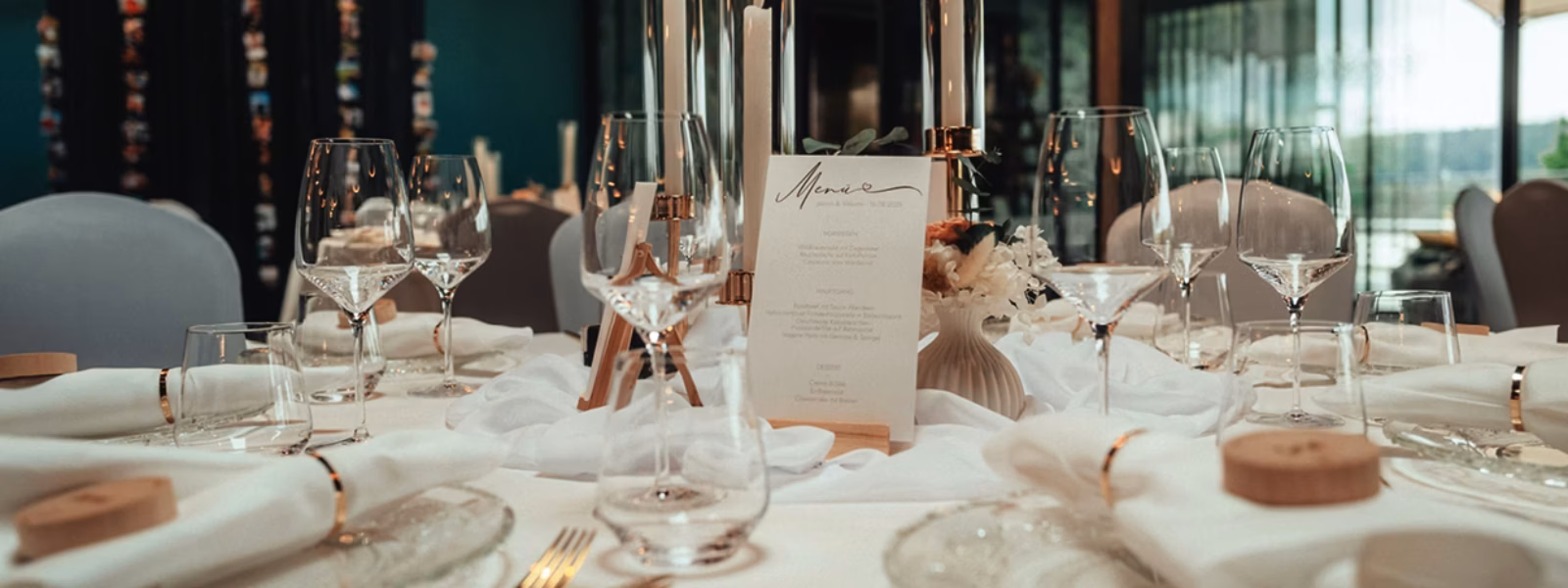 Nahaufnahme eines elegant gedeckten Tisches mit Gläsern, weißen Tellern, goldenem Besteck, weißen Servietten, hohen Kerzen und einer gedruckten Speisekarte als Tafelaufsatz in einem modernen, gut beleuchteten Lokal.