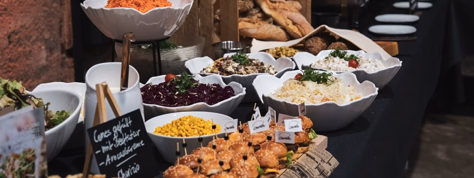 Ein Buffettisch mit verschiedenen Gerichten in weißen Schüsseln, darunter Salate, Mais und geschreddertes Gemüse, daneben Schieber, Brot und beschriftete Lebensmittelschilder. Im Hintergrund sind Teller und Utensilien aufgestellt.