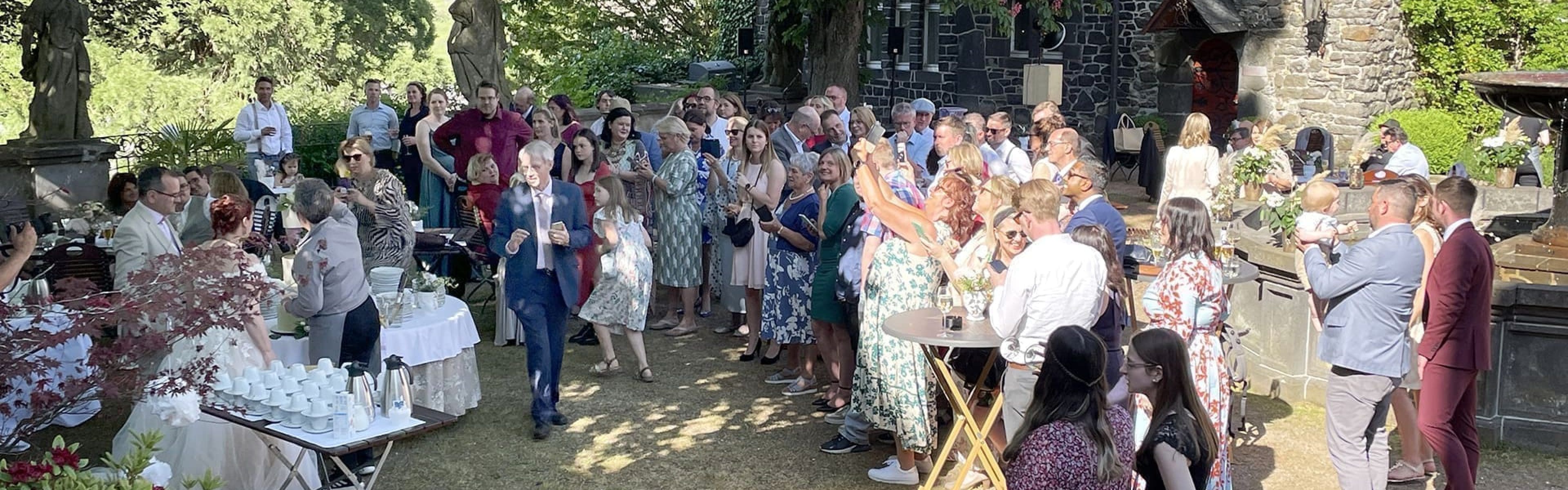 Eine große Gruppe von Menschen versammelt sich im Freien in der Nähe eines Steingebäudes, einige stehen, andere sitzen an Tischen. Die Szene umfasst Bäume, Statuen und Erfrischungen auf Tischen, was auf ein gesellschaftliches Ereignis oder eine Feier an einem sonnigen Tag hindeutet.