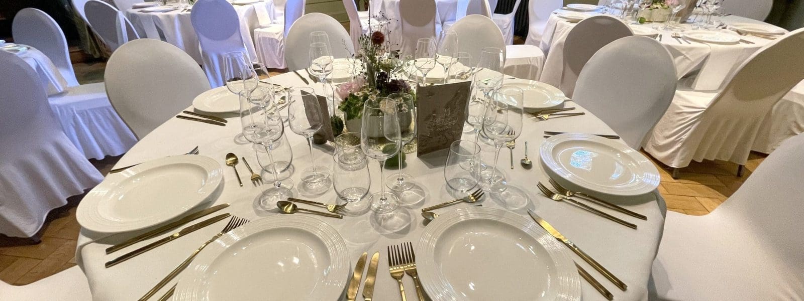 Ein runder, für sechs Personen gedeckter Esstisch mit weißen Tellern, goldenem Besteck, Kristallgläsern und einem blumengeschmückten Tafelaufsatz in einem eleganten Raum; im Hintergrund stehen weitere ähnlich gedeckte Tische mit weißen Stuhlhussen.