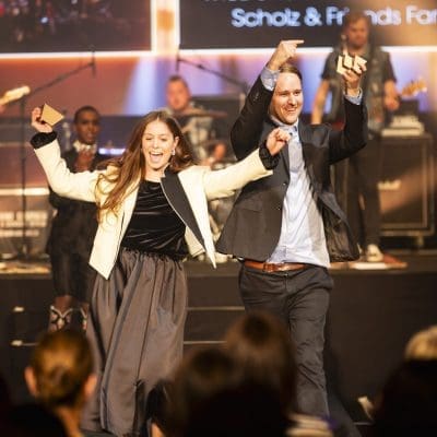 Ein Mann und eine Frau in förmlicher Kleidung gehen energisch mit erhobenen Armen auf die Bühne und lächeln. Im Hintergrund spielt eine Live-Band im Bühnenlicht. Im Vordergrund ist das Publikum zu sehen.