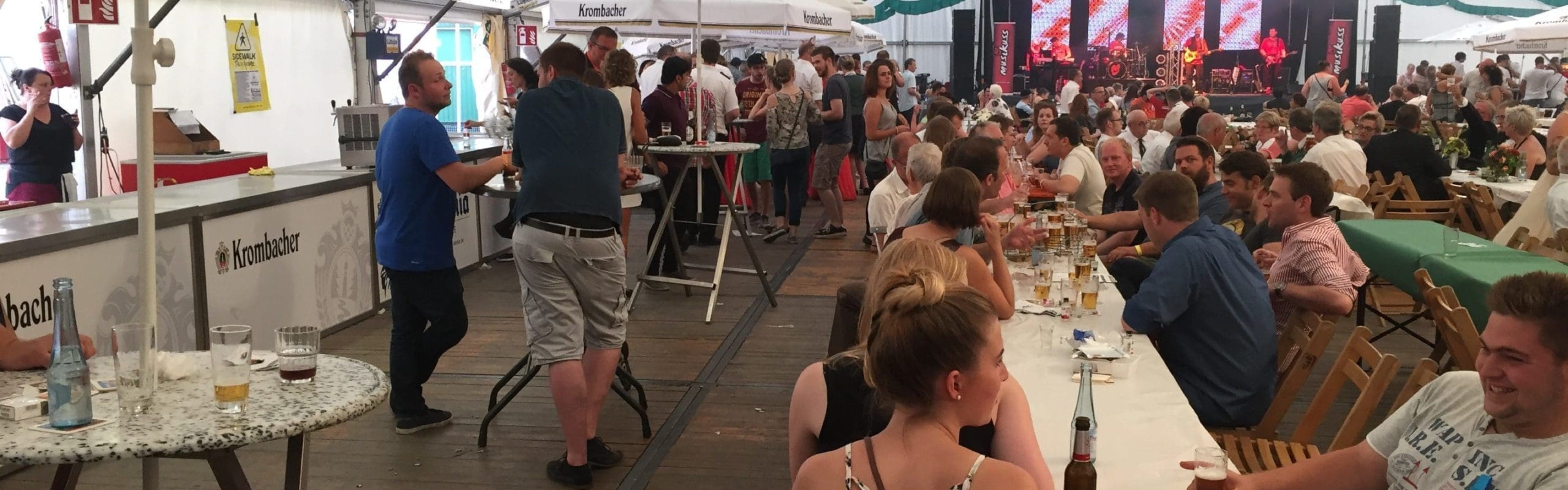 Die Menschen sitzen an langen Tischen in einem großen Zelt, trinken Bier und unterhalten sich. Einige stehen in der Nähe des Barbereichs. Das Zelt ist mit grünen Bannern geschmückt, ein Regenschirm wirbt für Krombacher, und im Hintergrund ist eine Bühne mit Lichtern zu sehen.