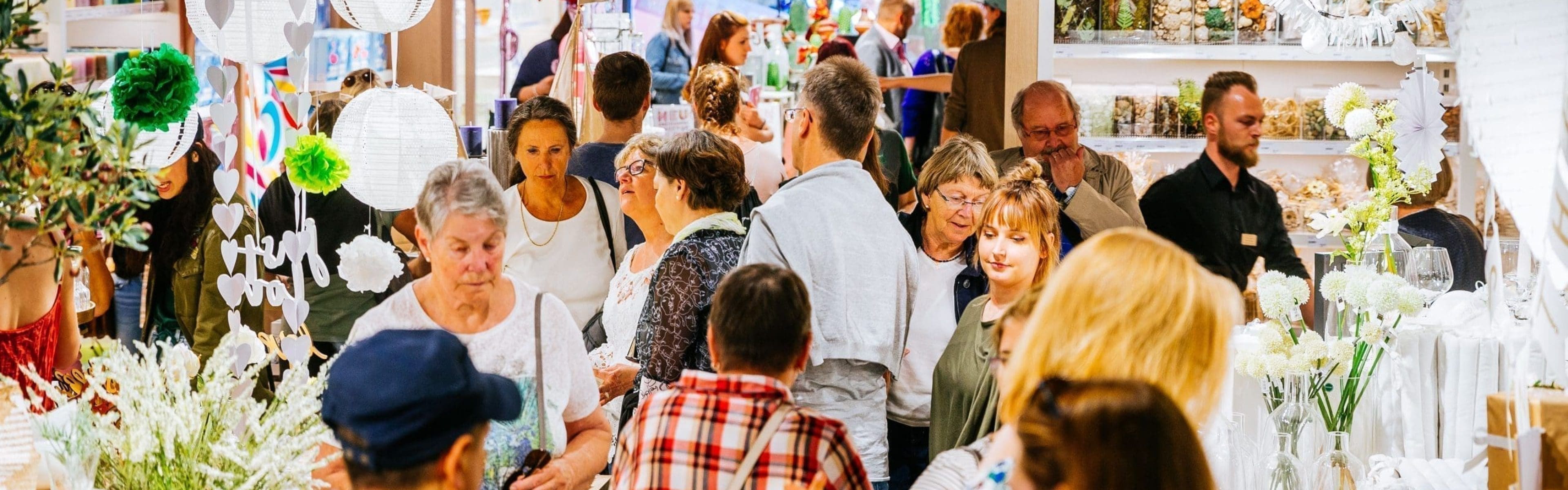 Ein überfüllter Geschenkartikelladen, in dem die Leute in verschiedenen Regalen und Auslagen mit Dekoartikeln, Papierlaternen und Kunstblumen stöbern. Der Laden ist hell erleuchtet und geschäftig, und die Kunden bewegen sich durch die engen Gänge.