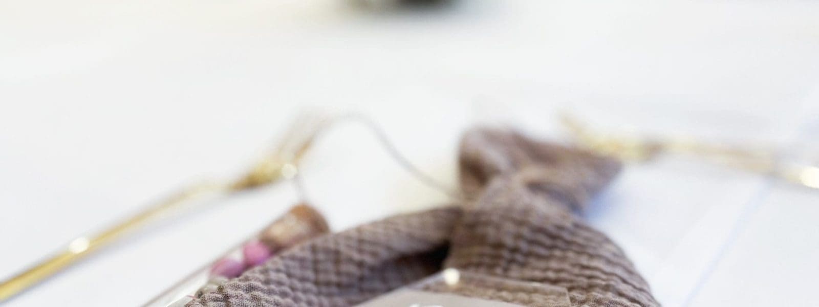 Eine strukturierte taupefarbene Serviette ist auf einer weißen Tischdecke mit einer sechseckigen durchsichtigen Tischkarte mit der Aufschrift Braut gefaltet. Im Hintergrund sind unscharfes goldenes Besteck und Weichzeichnerdekor zu sehen.