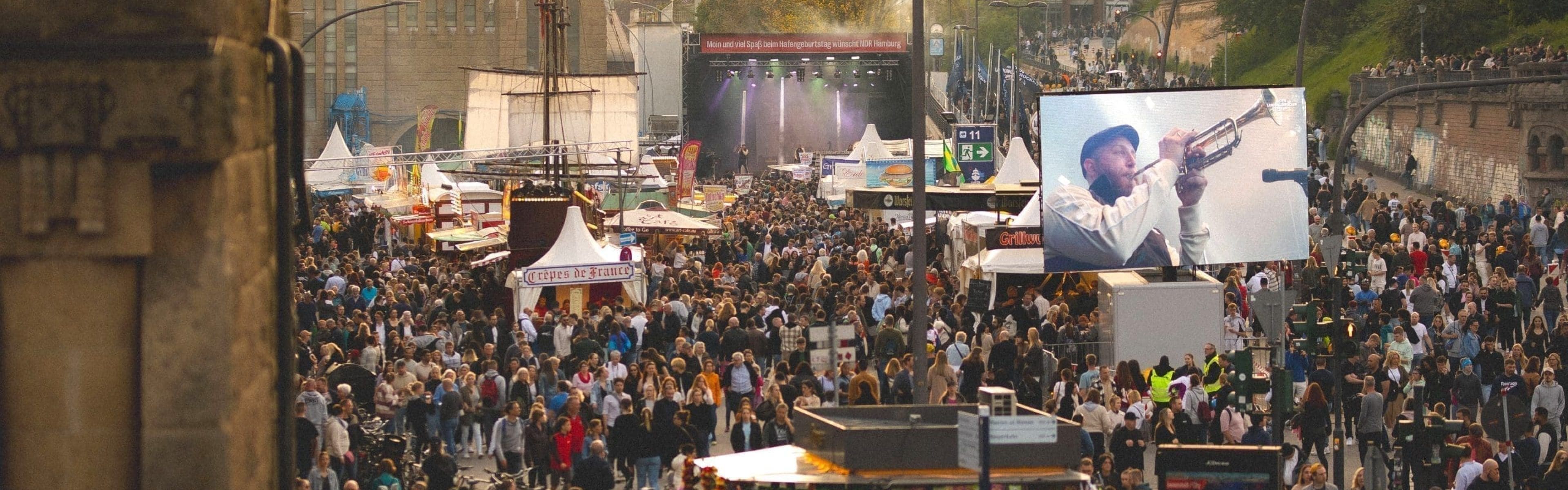 Eine große Menschenmenge versammelt sich bei einem Fest im Freien mit Essensständen und einer Bühne im Hintergrund. Ein Bildschirm zeigt eine Nahaufnahme eines Trompete spielenden Musikers. In der Ferne sind Gebäude und Bäume zu sehen.