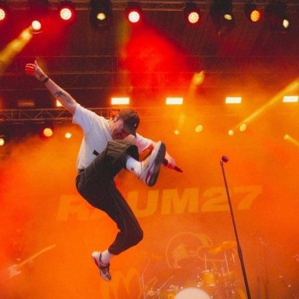 Ein Musiker in weißem Hemd und schwarzer Hose springt mit einem Mikrofon in der Hand über die Bühne, während im Hintergrund orangefarbene Bühnenlichter und Rauch zu sehen sind und eine Band spielt.