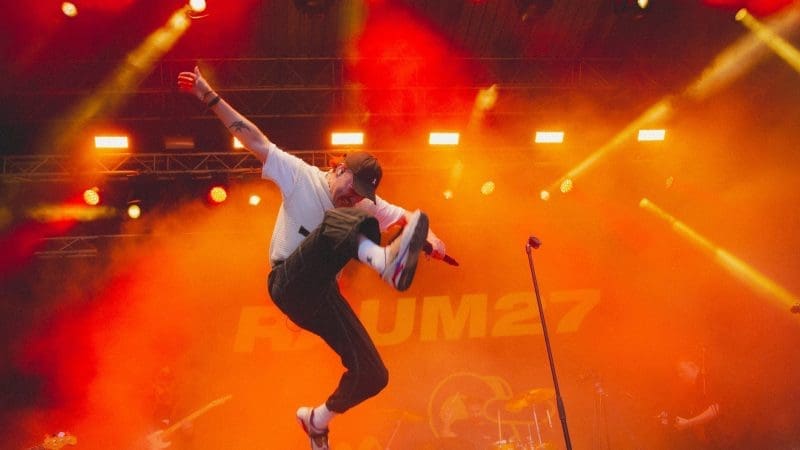 Ein Musiker in weißem Hemd und schwarzer Hose springt mit einem Mikrofon in der Hand über die Bühne, während im Hintergrund orangefarbene Bühnenlichter und Rauch zu sehen sind und eine Band spielt.