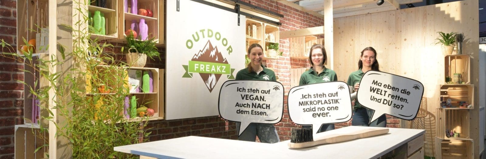 Drei Personen stehen in einem Holzstand mit Outdoor Freakz-Schildern und halten Sprechblasen mit deutschem Text über Veganismus, Vermeidung von Mikroplastik und Umweltschutz. In Regalen stehen Flaschen und Pflanzen.