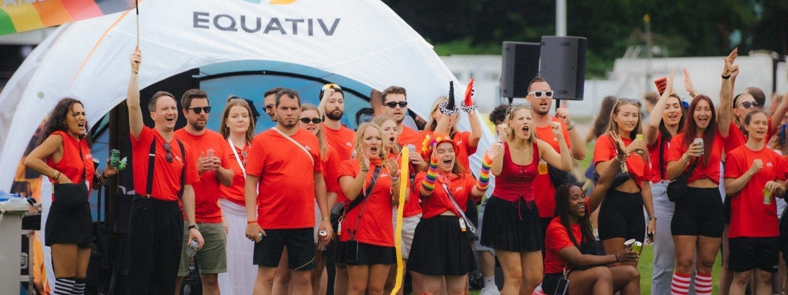 Eine Gruppe von Menschen in roten Hemden und mit bunten Accessoires steht jubelnd auf dem Rasen vor einem weißen Zelt mit dem Equativ-Logo bei einer Freiluftveranstaltung. Einige halten Getränke, Pom Poms und eine Flagge in der Hand.