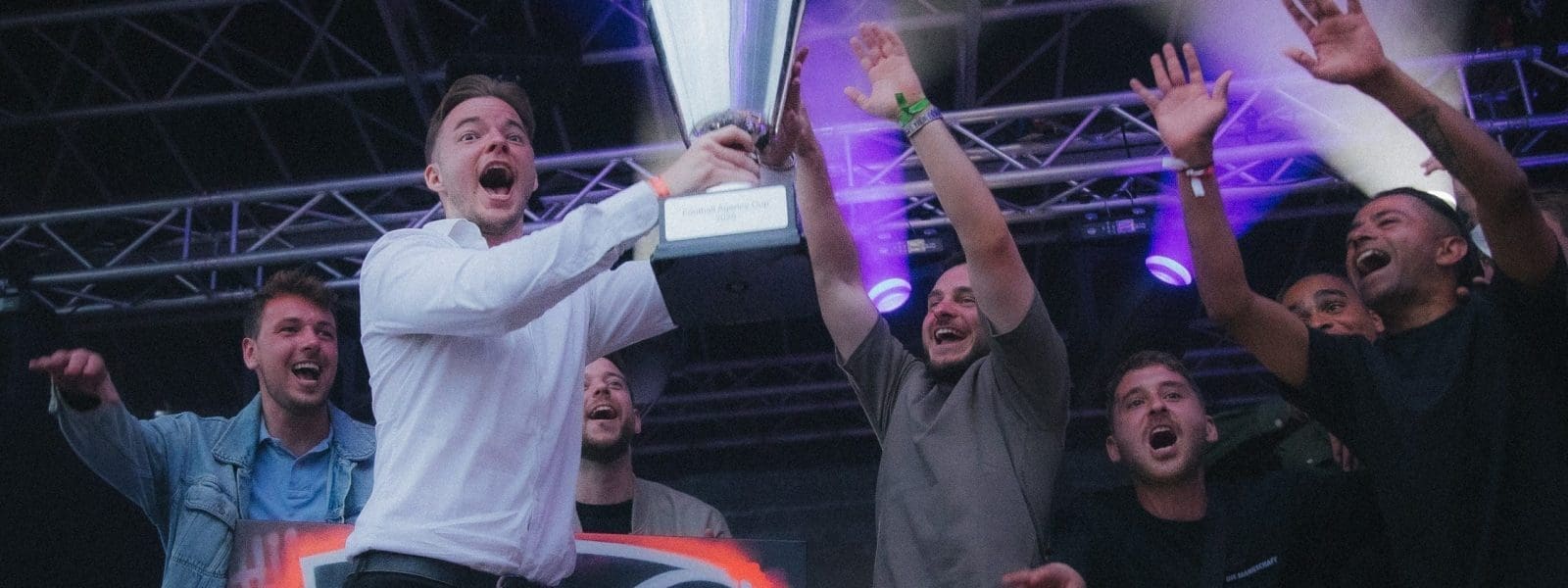 Eine Gruppe von Menschen auf einer Bühne feiert, wobei eine Person in einem weißen Hemd einen großen Pokal hochhält, während andere jubeln und die Hände heben. Im Hintergrund sind Bühnenbeleuchtung und eine Metalltraverse zu sehen.