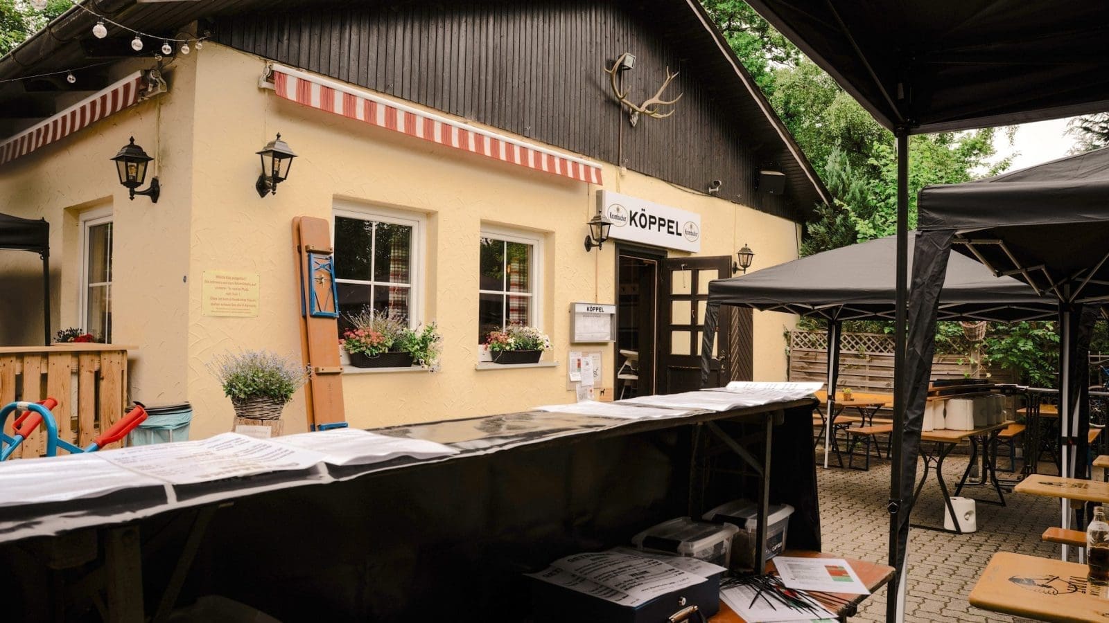 Außensitzbereich eines kleinen Restaurants oder Cafés namens Köppel mit Holztischen und -bänken, einem cremefarbenen Gebäude, Topfpflanzen und einer gestreiften Markise, die unter einer Baumkrone sichtbar ist.