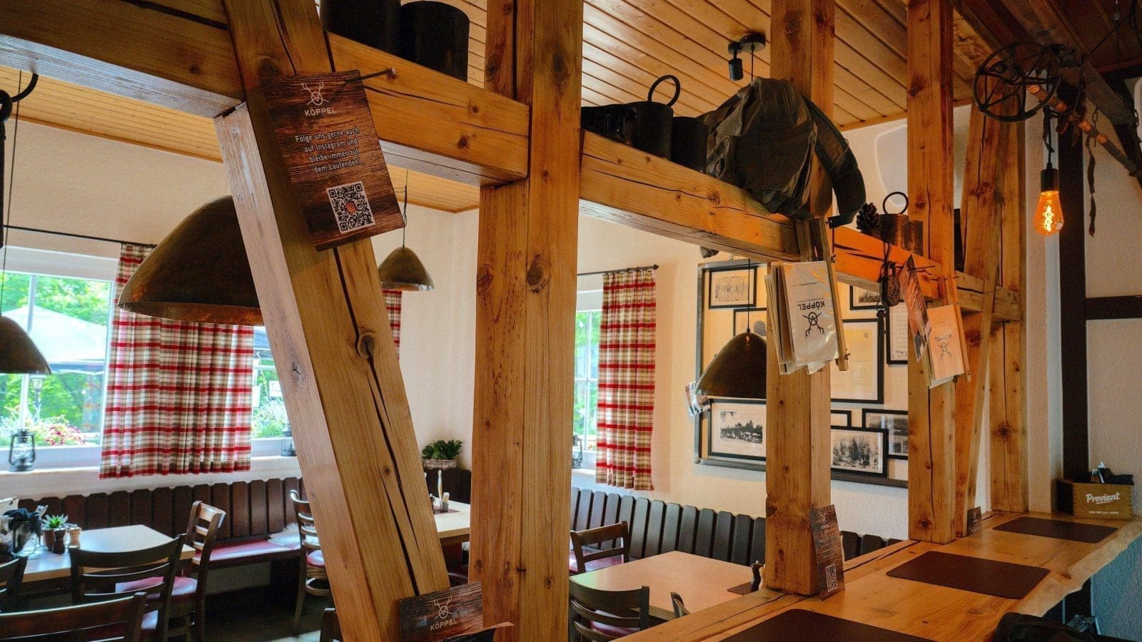 Ein gemütliches Restaurant mit Holzbalken, Tischen mit braunen Tischsets, rot-weiß karierten Vorhängen, gerahmten Bildern an den Wänden und Hängelampen an der Decke.