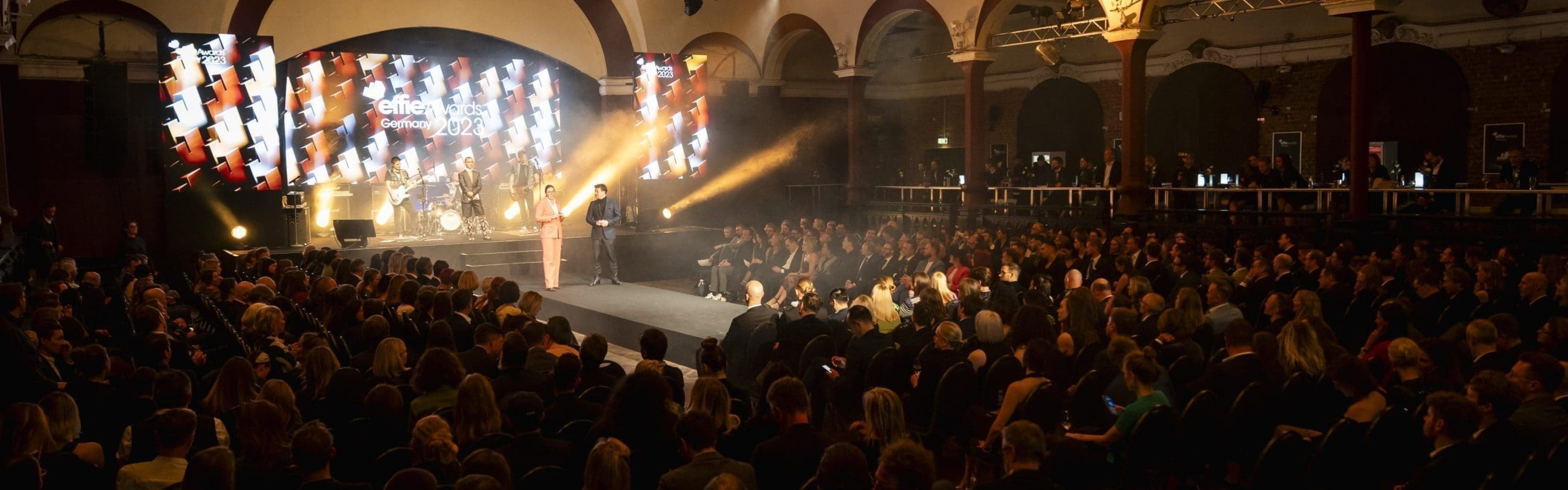 Eine große Menschenmenge sitzt vor einer Bühne, auf der eine Person unter hellem Licht spricht. Der Veranstaltungsort hat gewölbte Decken, zwei große Bildschirme auf beiden Seiten der Bühne und Scheinwerfer, die die Szene beleuchten.