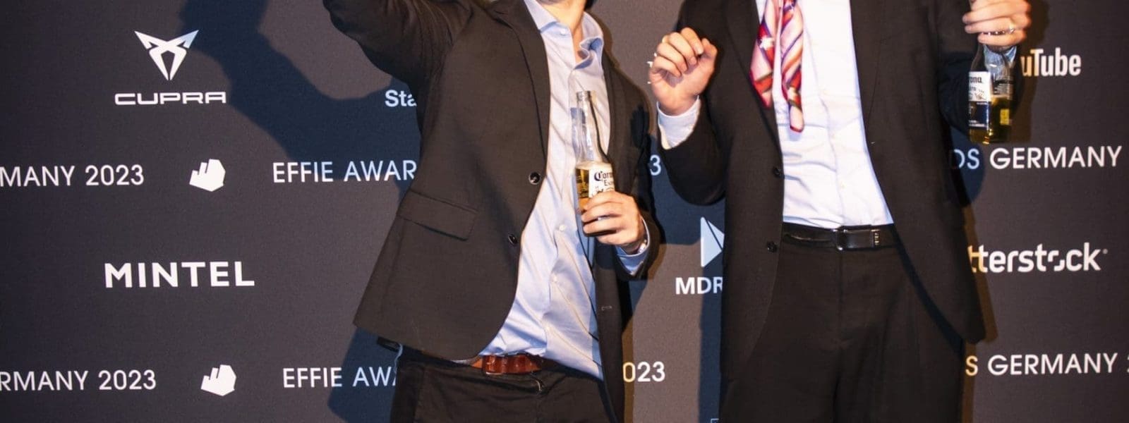 Zwei Männer in Anzügen stehen auf einem schwarzen Teppich vor einer Kulisse mit dem Schriftzug Effie Awards Germany 2023. Sie lächeln, halten Drinks in der Hand, und ein Mann hebt feierlich den Arm.