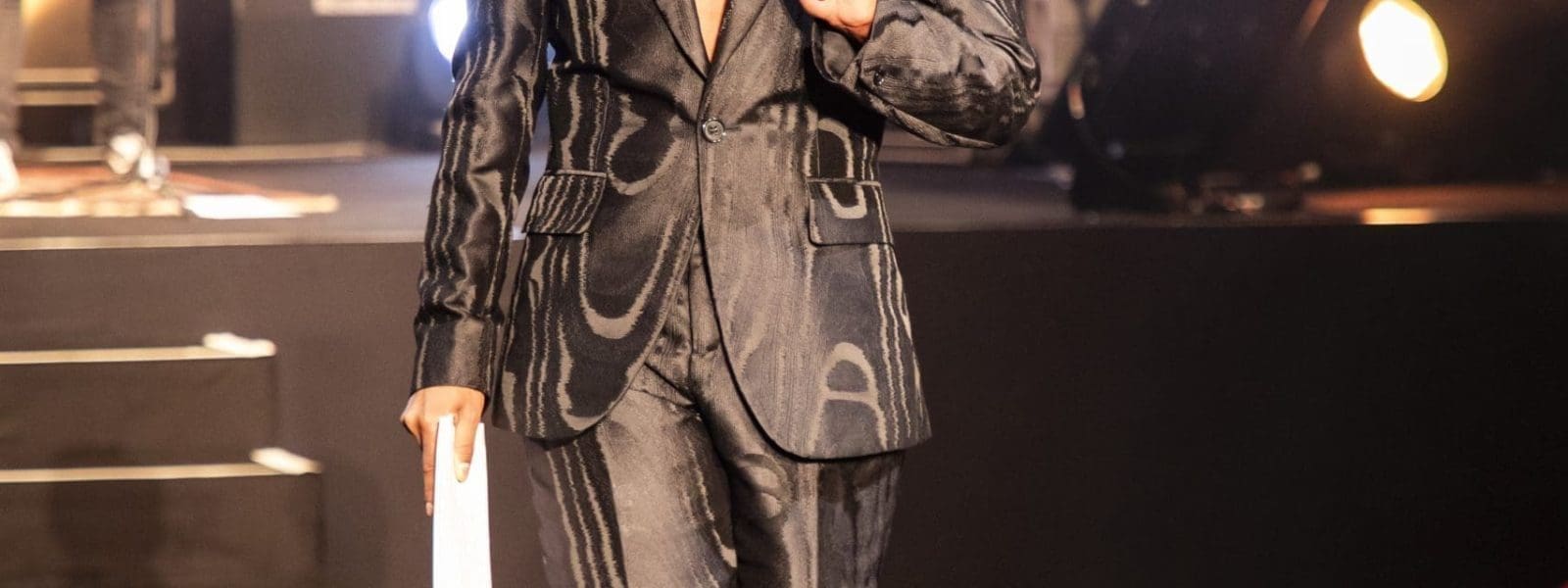 Eine Person geht einen Laufsteg hinunter und trägt eine dunkle, glänzende Anzugjacke, dazu passende knielange Shorts, gemusterte kniehohe Stiefel und ein juwelenbesetztes Halsband. Im Hintergrund sind Bühnenlichter und eine Gitarre spielende Person zu sehen.