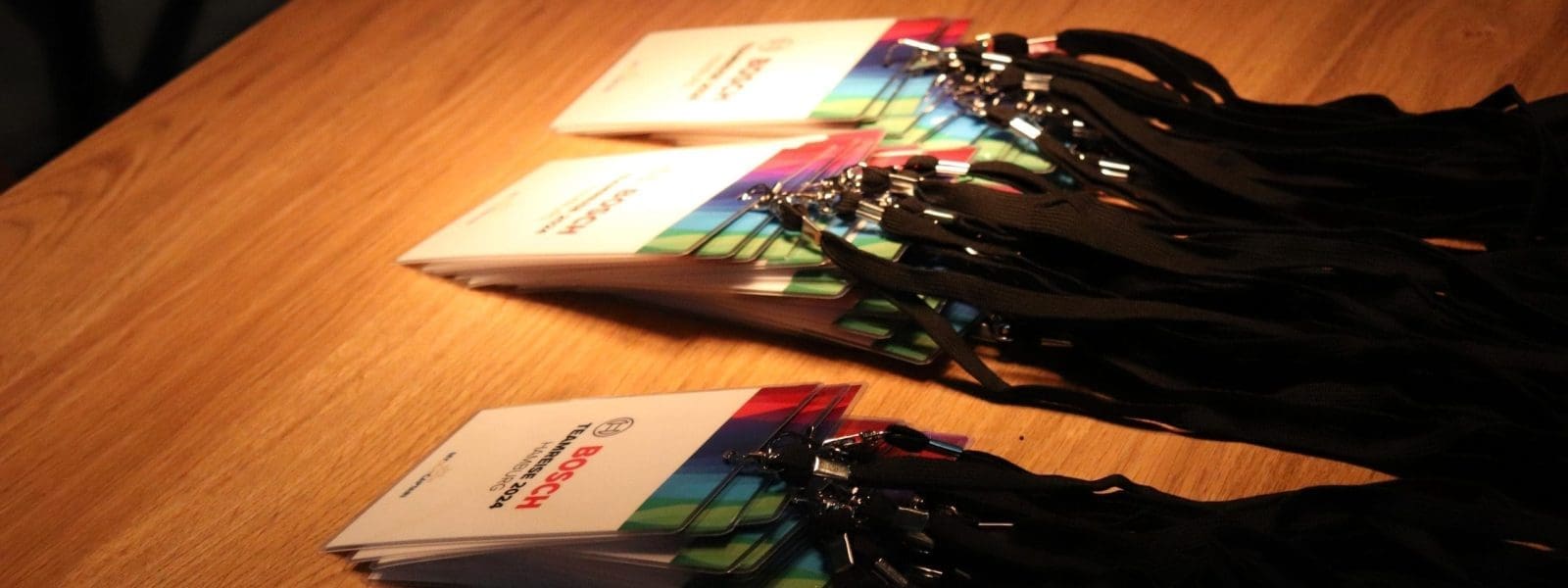 Auf einem Holztisch liegen mehrere Stapel von Veranstaltungsplaketten mit schwarzen Lanyards, die von einem Oberlicht beleuchtet werden. Auf den Ansteckern sind Text und ein Bosch-Logo abgebildet.