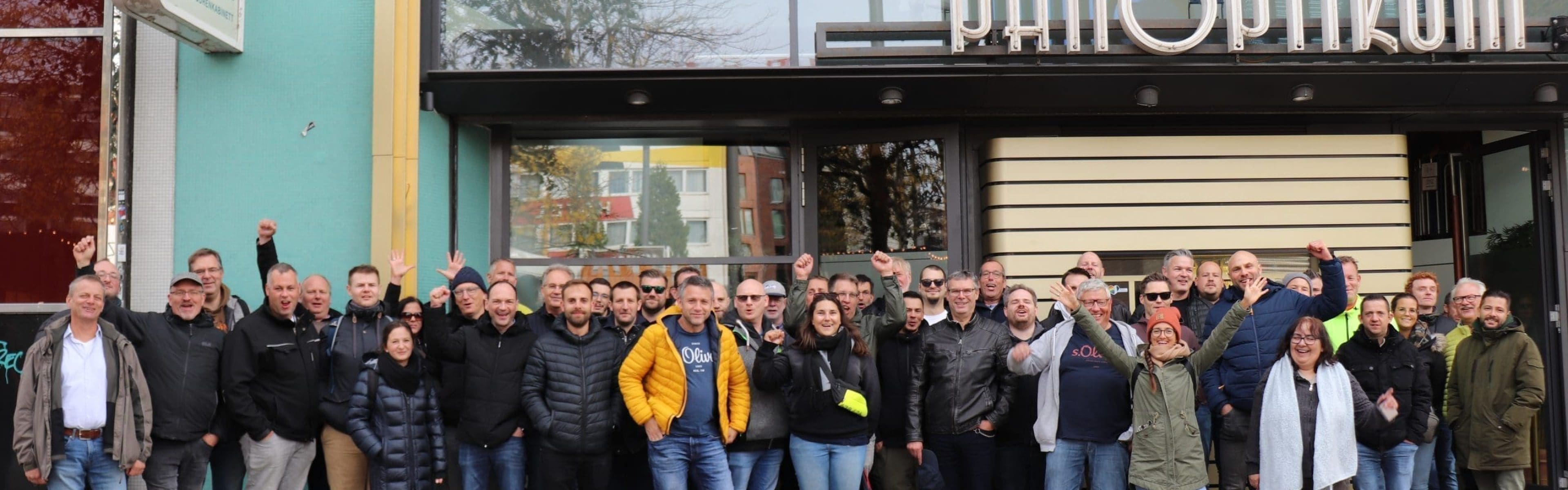 Eine große Gruppe von Menschen steht vor dem Eingang des Panoptikum-Museums und posiert für ein Foto; einige lächeln und heben die Arme. Das Gebäude hat große Fenster und ein Schild mit "Panoptikum" über der Tür.