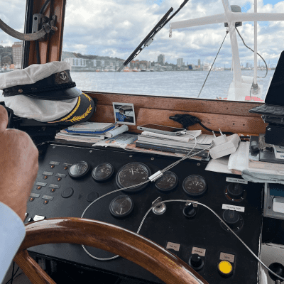 Blick aus dem Steuerhaus eines Schiffes mit Steuerrad, Navigationsinstrumenten, einer Kapitänsmütze auf dem Armaturenbrett, verschiedenen Papieren und Geräten sowie einer Hand, die das Steuerrad ergreift, wobei das Wasser und die Skyline der Stadt durch das Fenster zu sehen sind.