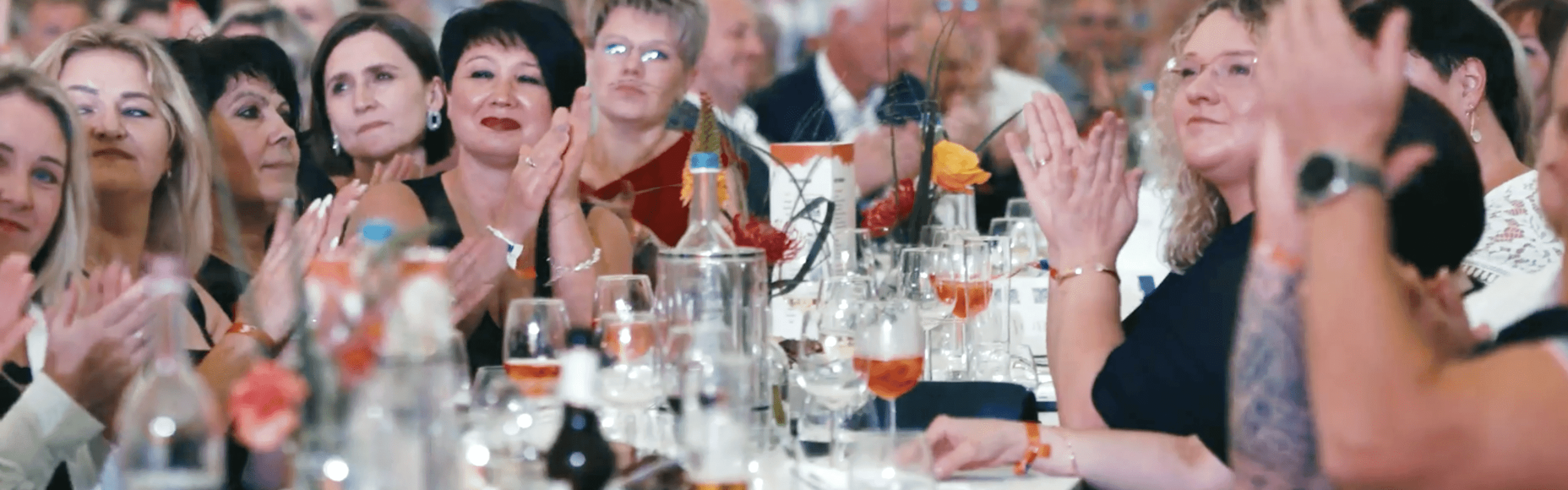 Eine große Gruppe von Menschen sitzt an einem langen Tisch mit Getränken und Blumen, klatschend und nach vorne gewandt, in einem hellen, überfüllten Innenraum.