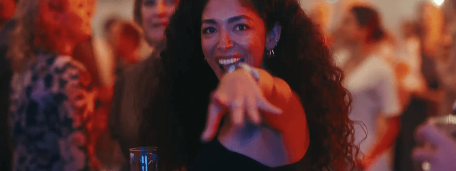 Eine Frau mit langen lockigen Haaren lächelt und zeigt in die Kamera, während sie ein Getränk in der Hand hält, umgeben von einer unscharfen Menschenmenge bei einer lebhaften Veranstaltung in einer Halle.