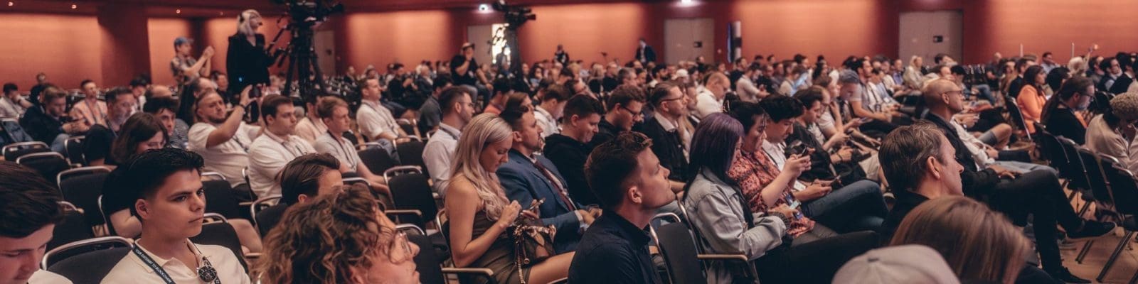 Ein großes Publikum sitzt in Stuhlreihen bei einer Konferenz oder Veranstaltung in einer Halle, wobei viele Leute nach vorne schauen und einige elektronische Geräte benutzen. An der Decke sind rechteckige Hängelampen angebracht.