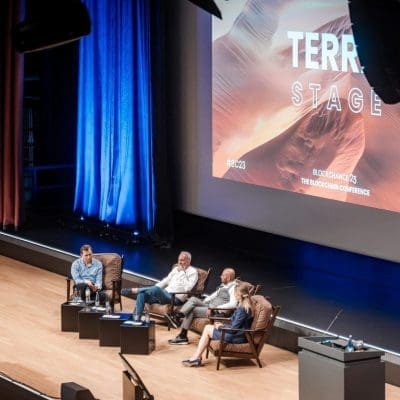 Drei Personen sitzen auf einer Bühne und führen eine Podiumsdiskussion vor einem Publikum. Auf einer großen Leinwand hinter ihnen steht TERRA STAGE. Die Bühne ist mit blauem und orangefarbenem Licht beleuchtet. Eine Kamera filmt von vorne.