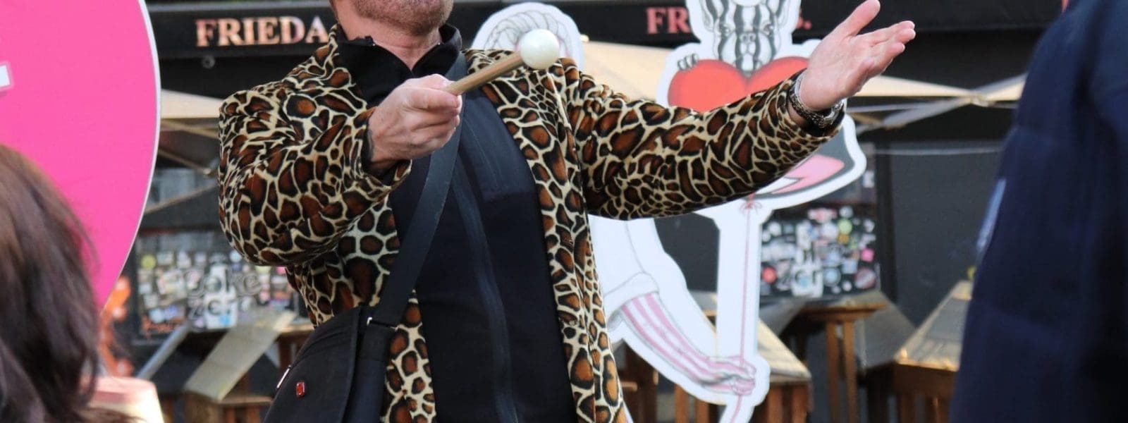 Eine Person, die einen Mantel mit Leopardenmuster und einen Hut trägt, gestikuliert mit einem erhobenen Arm, während sie im Freien zu einer kleinen Menschenmenge spricht. Im Hintergrund sind Regenbogenfahnen und dekorierte Schaufenster zu sehen.