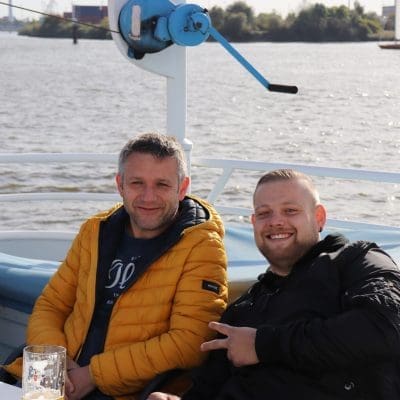 Zwei Männer sitzen Seite an Seite auf einem Boot mit einem Fluss im Hintergrund. Der eine trägt eine gelbe Jacke, der andere eine schwarze Jacke, lächelt und macht ein Friedenszeichen. Auf dem Tisch stehen Biergläser und eine Schachtel Zigaretten.