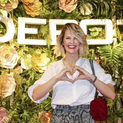 Eine Person mit blondem Haar und rotem Lippenstift steht vor einem Hintergrund aus Blumen und Grünpflanzen, auf dem in großen weißen Buchstaben DEPOT geschrieben steht. Sie lächelt und macht mit ihren Händen eine Herzform.