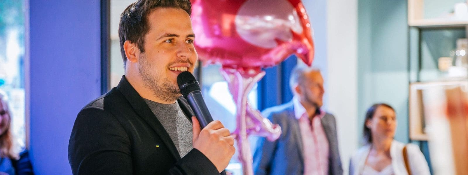 Ein Mann in einem schwarzen Blazer hält ein Mikrofon in der Hand und spricht in einem Raum. Hinter ihm ist ein rosafarbener Flamingo-Ballon zu sehen, und im Hintergrund stehen drei Personen, von denen eine zuzuhören scheint.