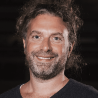 Eric Ein Mann mit lockigem braunem Haar und Bart lächelt in die Kamera. Er trägt ein dunkles Hemd mit V-Ausschnitt und ist von den Schultern aufwärts vor einem dunklen Hintergrund zu sehen.