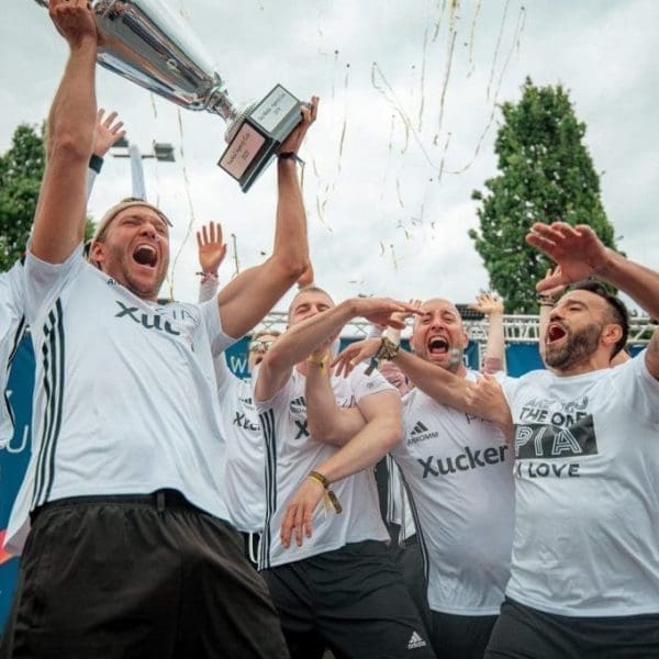 Eine Gruppe von Männern in weißen Fußballtrikots feiert, während einer einen Pokal hochhält. Konfetti fällt in die Luft und einige Spieler heben lächelnd und jubelnd die Arme. Im Hintergrund sind Bäume und eine blaue Fahne zu sehen.