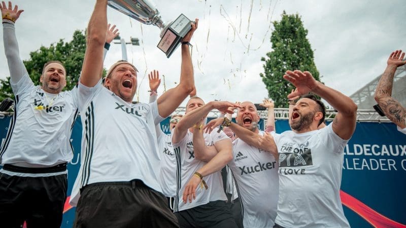 Eine Gruppe von Männern in weißen Fußballtrikots feiert, während einer einen Pokal hochhält. Konfetti fällt in die Luft und einige Spieler heben lächelnd und jubelnd die Arme. Im Hintergrund sind Bäume und eine blaue Fahne zu sehen.