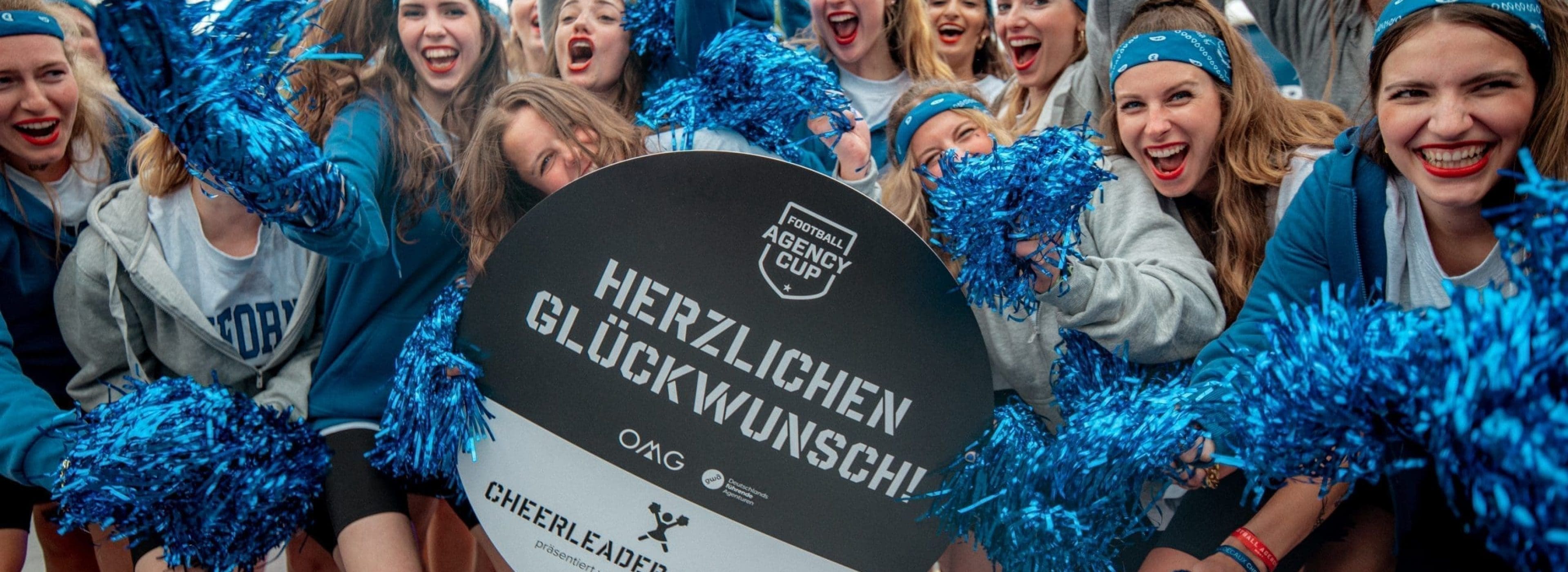Eine Gruppe lächelnder Cheerleader in blauen Outfits und mit blauen Pom-Poms posiert im Freien und hält ein großes rundes Schild mit der Aufschrift HERZLICHEN GLÜCKWUNSCH! CHEERLEADER-CUP 2023.