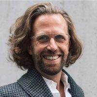 Fabian_10x10 Ein Mann mit schulterlangem braunem Haar, Bart und Brille lächelt. Er trägt einen karierten Mantel über einem weißen Hemd und steht vor einer hellen, strukturierten Wand.